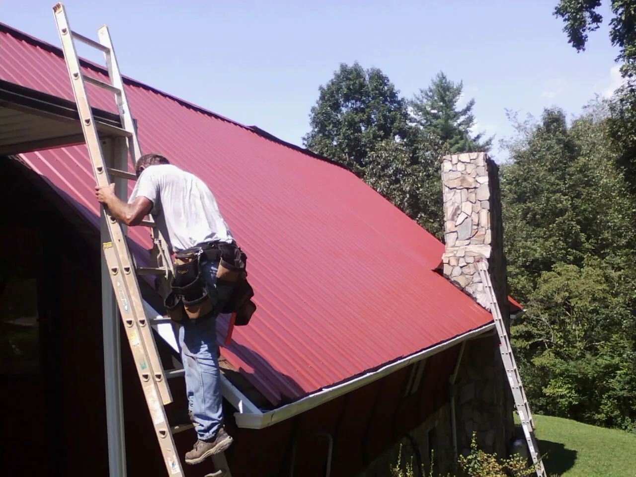 Roofer on a ladder beside a red metal roof and stone chimney — Soffit & Fascia Repair by Roofing Brothers in Cooksville